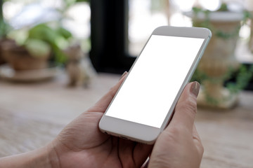 Mockup image of woman's hands holding mobile phone with blank white screen on wooden table in vintage cafe