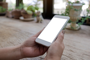 Mockup image of woman's hands holding mobile phone with blank white screen on wooden table in vintage cafe