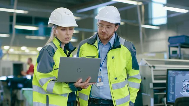 Male and Female Industrial Engineers Wearing safety,  Work on a Factory, They Discuss Project while Using Laptop.In the Background Industrial Facility.