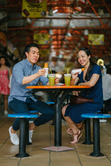 A portrait of a young Chinese couple spending time together on a date. Both are drinking sugarcane juice and eating fried bananas.