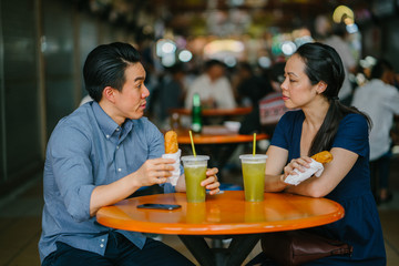 An image of a young Asian Chinese couple on their date at a hawker center. Both are enjoying their fried bananas and sugarcane juice.