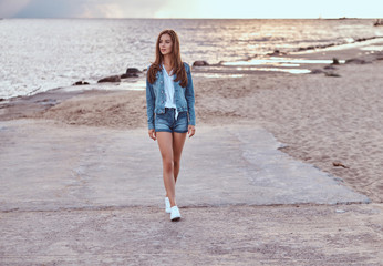 Fototapeta na wymiar Full body portrait of a sensual girl dressed in denim shorts and jacket walks on the beach in cloudy weather during sunset.
