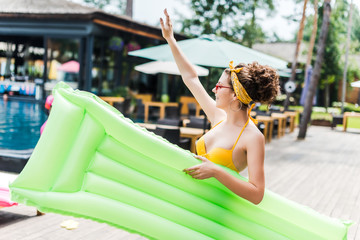 girl in swimsuit walking with inflatable mattress near swimming pool and waving hand