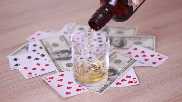 Pouring glass with whisky beside playing cards and dollar banknotes