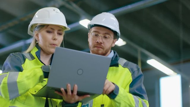 Male and Female Industrial Engineers Work on a Manufacturing Plant, They Discuss Project, Point in the Direction of the Machinery while Using Laptop. In the Background Industrial Manufacturing Plant.