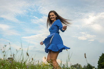 Beautiful smiling young woman is dancing and whirls in a blue dress in a meadow with a flowers at flowers background