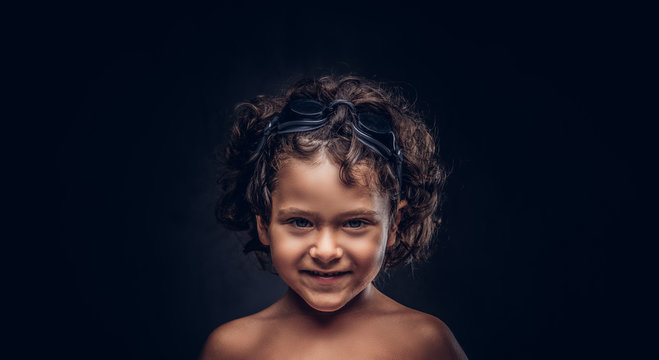 Smiling little shirtless boy in swimming goggles posing in a studio. Isolated on the dark textured background.
