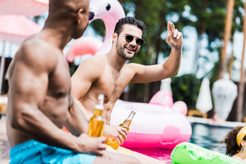 smiling man in sunglasses gesturing by hand while his friend sitting near with beer on poolside