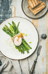 Healthy breakfast. Flat-lay of green asparagus with soft-boiled egg, leek and bacon served in white plate and grilled bread slices over rustic wooden background, top view