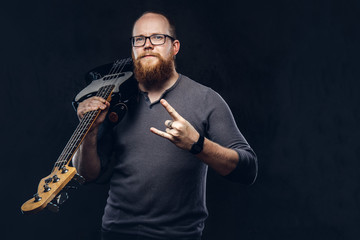 Redhead bearded male musician wearing glasses dressed in a gray t-shirt holds electric guitar and show rock and roll sign. Isolated on a dark textured background.