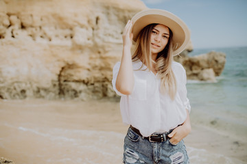 Young beautiful woman wearing straw hat posing standing on beach near waves and rock and looking away at the sea