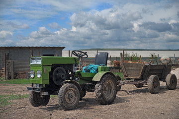 A small green tractor stands on the farm in a hook with a cart