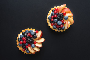 Short-pastry sweet baked tartlets with fresh ripe berries and fruits on black background view...
