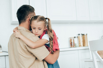 father and sad little daughter with backpack hugging each other in kitchen at home, back to school...