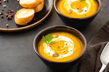 Pumpkin soup in a bowl with fresh pumpkins, garlic and parsley herbs on a black background. Autumn concept.