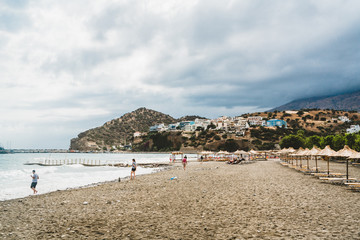 Fototapeta premium Crete, Greece. View from cliffs to village with marine vessels, boats and lighthouse. View from cliff on Bay with beach and architecture - vacation destination resort with clear ocean water. Rethymno