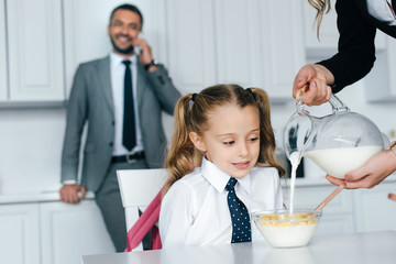 selective focus of kid in school uniform at table with breakfast and mother pouring milk into bowl with crunches at home, back to school concept