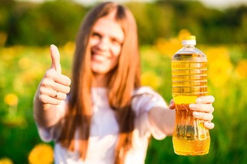 Young woman in white holding bottle of oil in sunflower field and show thumbs up