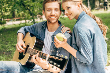 Fototapeta premium handsome young man playing guitar for his smiling girlfriend and looking at camera while having picnic at park