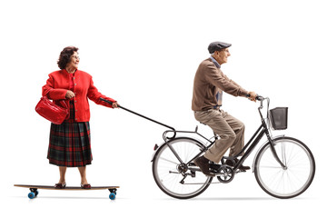 Elderly man on a bicycle pulling an elderly woman on a longboard