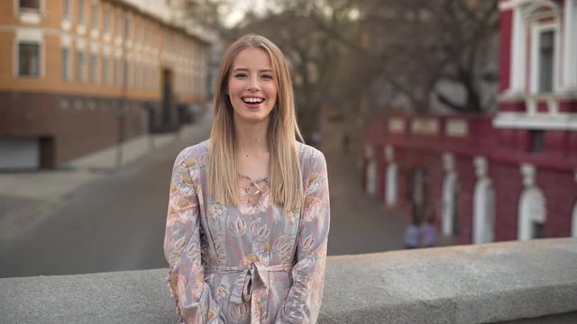 Portrait of seductive cheerful woman 20s with blond hair wearing dress laughing, while standing on bridge against city street slow motion