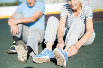 Mid section portrait of white haired senior couple tying sports shoes sitting on running track outdoors, copy space - Powered by Adobe
