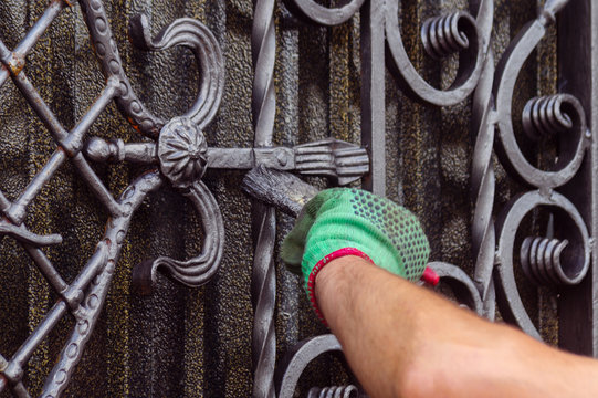 A man paints a gate with forged elements. On his hand is a green glove, near the elbow of a graze. The hand is located on the right of the frame.