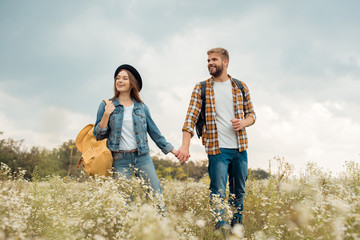 smiling couple with backpacks holding hands while walking in summer field with wild flowers