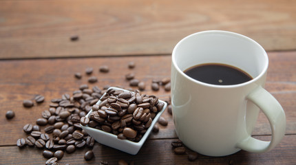 A cup of coffee with small white ceramic dish full of coffee beans on wooden background.
