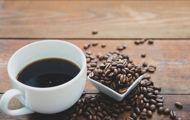 A cup of coffee with small white ceramic dish full of coffee beans on wooden background.