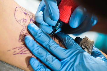 Master tattoo draws the black paint on the clients tattoo. Tattoo artist holding a tattoo machine in blue sterile gloves and working on the professional mat.