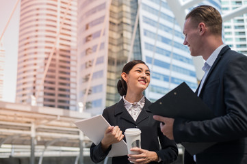 Asian business woman wear suit holding document file and plastic mug on hand walking together with  Caucasian businessman in the city and talk about business future.