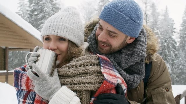 Loving young man smiling and embracing his beautiful girlfriend while she drinking hot tea from metal mug in forest at snowy winter day