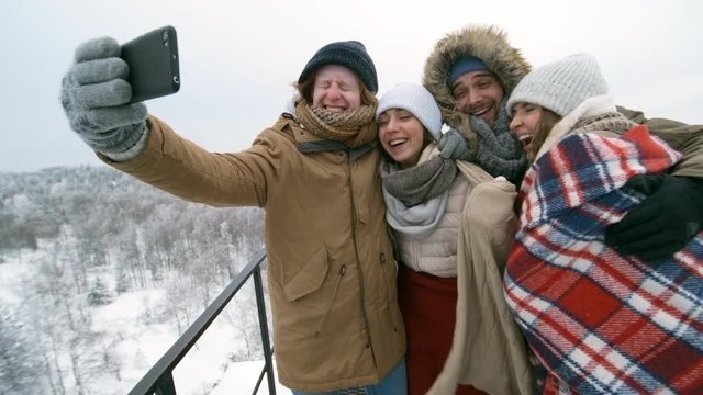 Group of young happy friends standing at viewing point in national park, smiling and hugging while taking selfie with smartphone at cold winter day