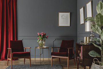 Two burgundy armchairs placed in grey living room interior with red drape. molding on the wall with posters, fresh flowers in glass vase and wooden cupboard