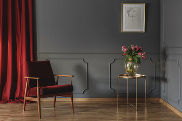 Red curtain and burgundy armchair standing in grey room interior with pink tulips on gold end table...