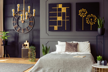 Plants and black and gold posters in grey bedroom interior with clock and bed with pillows. Real...
