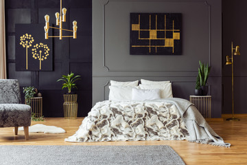 Black and gold poster on grey wall above bed in bedroom interior with plants and armchair. Real...