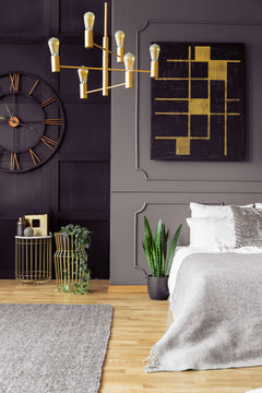 Plant next to bed in grey bedroom interior with chandelier, black poster and clock. Real photo