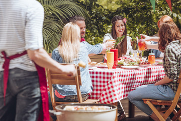 A group of young friends making a toast while sitting by a table full of food during a grill party outside.