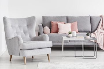 Comfy armchair and grey sofa with pink pillows, and coffee tables in a bright living room interior. Real photo
