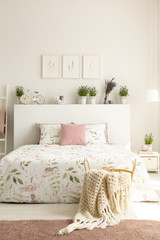 Real photo of basket with knit blanket standing by the bed with floral bedding in white bedroom interior with three simple posters, lavender plants and decor