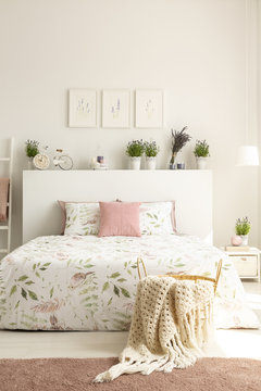 Real photo of basket with knit blanket standing by the bed with floral bedding in white bedroom interior with three simple posters, lavender plants and decor