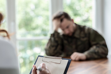 Close-up of a hand writing on a piece of paper with a depressed soldier in the blurred background