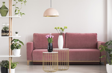 Real photo of a feminine living room interior with gold and marble tables with fresh flowers standing in front of a cozy pink sofa - obrazy, fototapety, plakaty