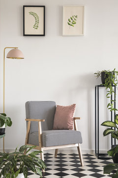 Pink and gold lamp standing next to a an armchair with houndstooth pattern in bright living room interior with many plants and pictures on white wall. Real photo