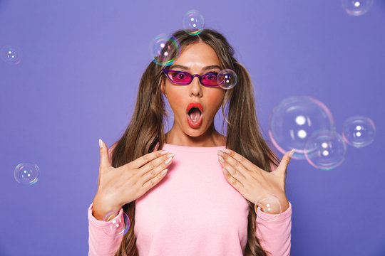 Portrait of excited teen woman with two ponytails in sweatshirt wearing trendy sunglasses wondering with open mouth, isolated over violet background in studio