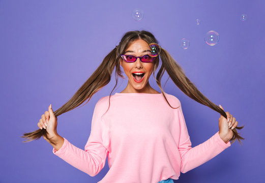 Photo of lovely caucasian woman in sweatshirt wearing trendy sunglasses having fun and touching her ponytails, isolated over violet background in studio