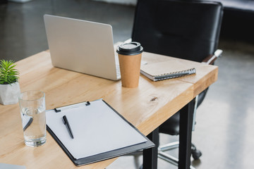 laptop, clipboard, glass of water and coffee to go on wooden table in office