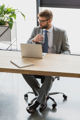 handsome young businessman in eyeglasses holding glass of water and using laptop